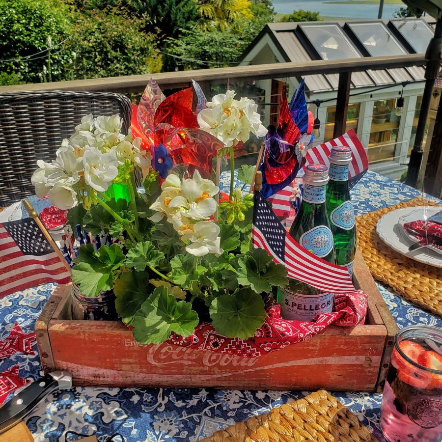 Patriotic centerpiece with red Coca Cola crate, white geraniums, American flags and pinwheels.