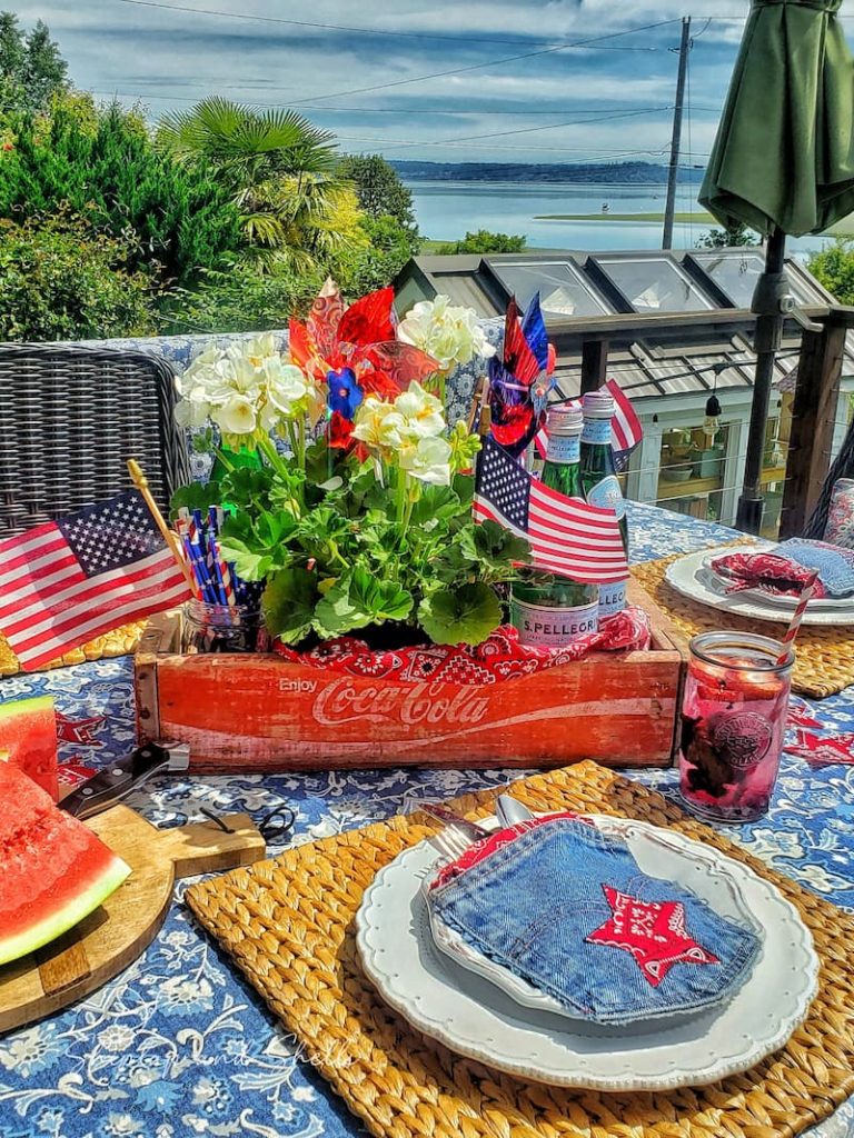 Patriotic centerpiece with red Coca Cola crate, white geraniums, American flags and pinwheels.