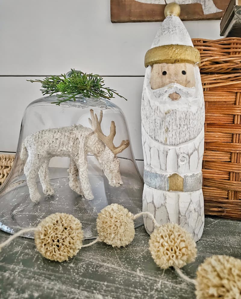 Green and white Christmas decor: white reindeer under glass cloche and white wooden Santa, and cream pom pom garland