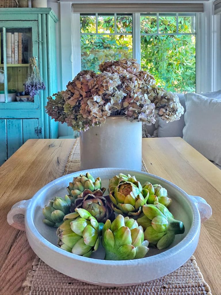 muted dried hydrangeas in a crock and a white ceramic bowl of artichokes on the kitchen table