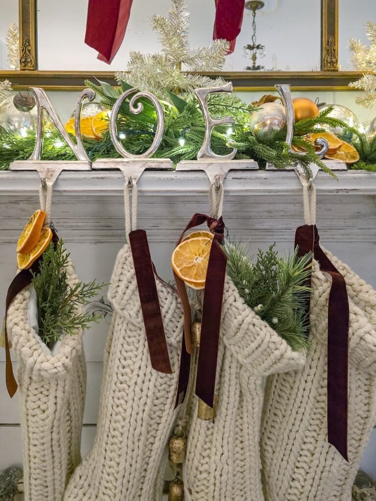 cream knit stockings hanging from silver NOEL hangers with brown velvet ribbon, greenery, bells, and dried oranges
