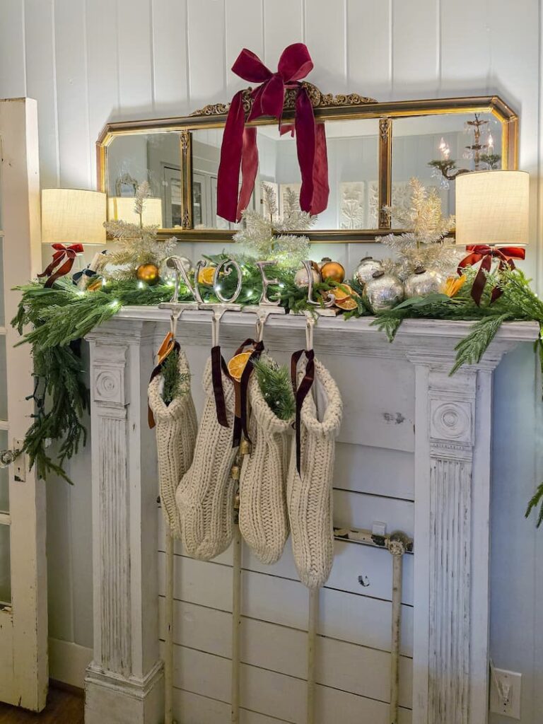 faux fireplace mantel decorated for Christmas with garland, cream knit stockings and a burgundy bow on mirror hanging above