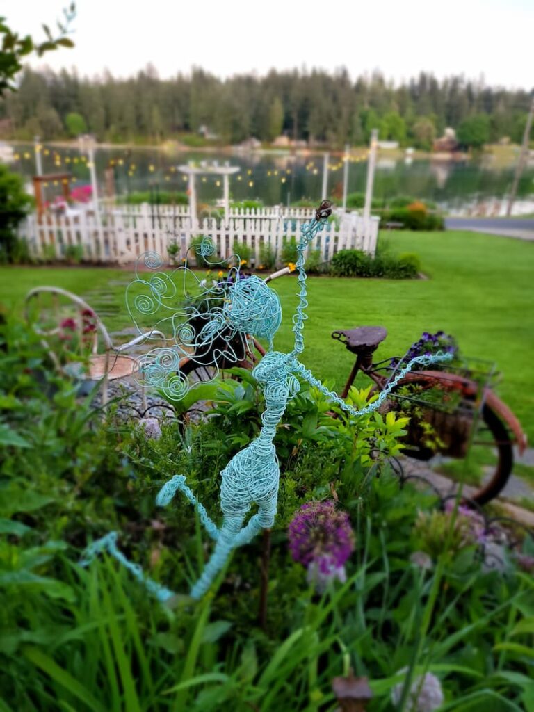 turquoise wire garden fairy in the cottage garden with vintage bike and white picket fence in the background