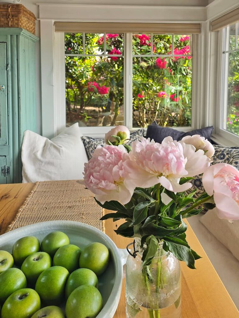 pale pink peonies in a vase, green apples in a bowl, and fuchsia rhododendrons growing in a tree outside the kitchen window