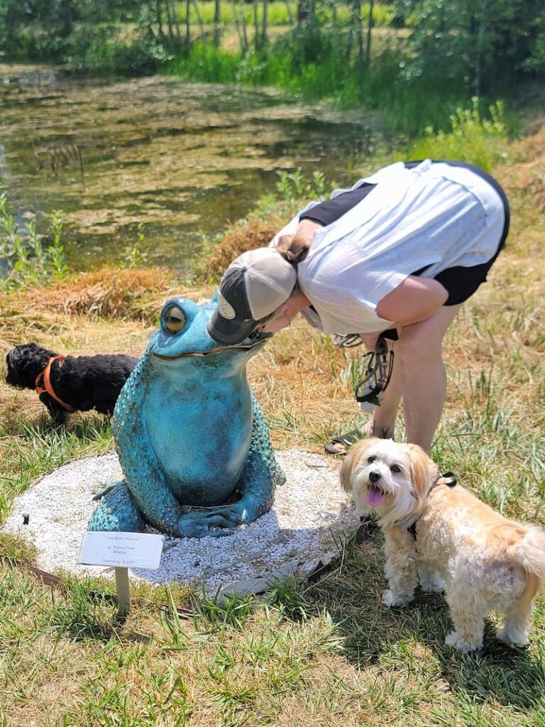 woman kissing a teal frog sculpture with black and cream dog looking on